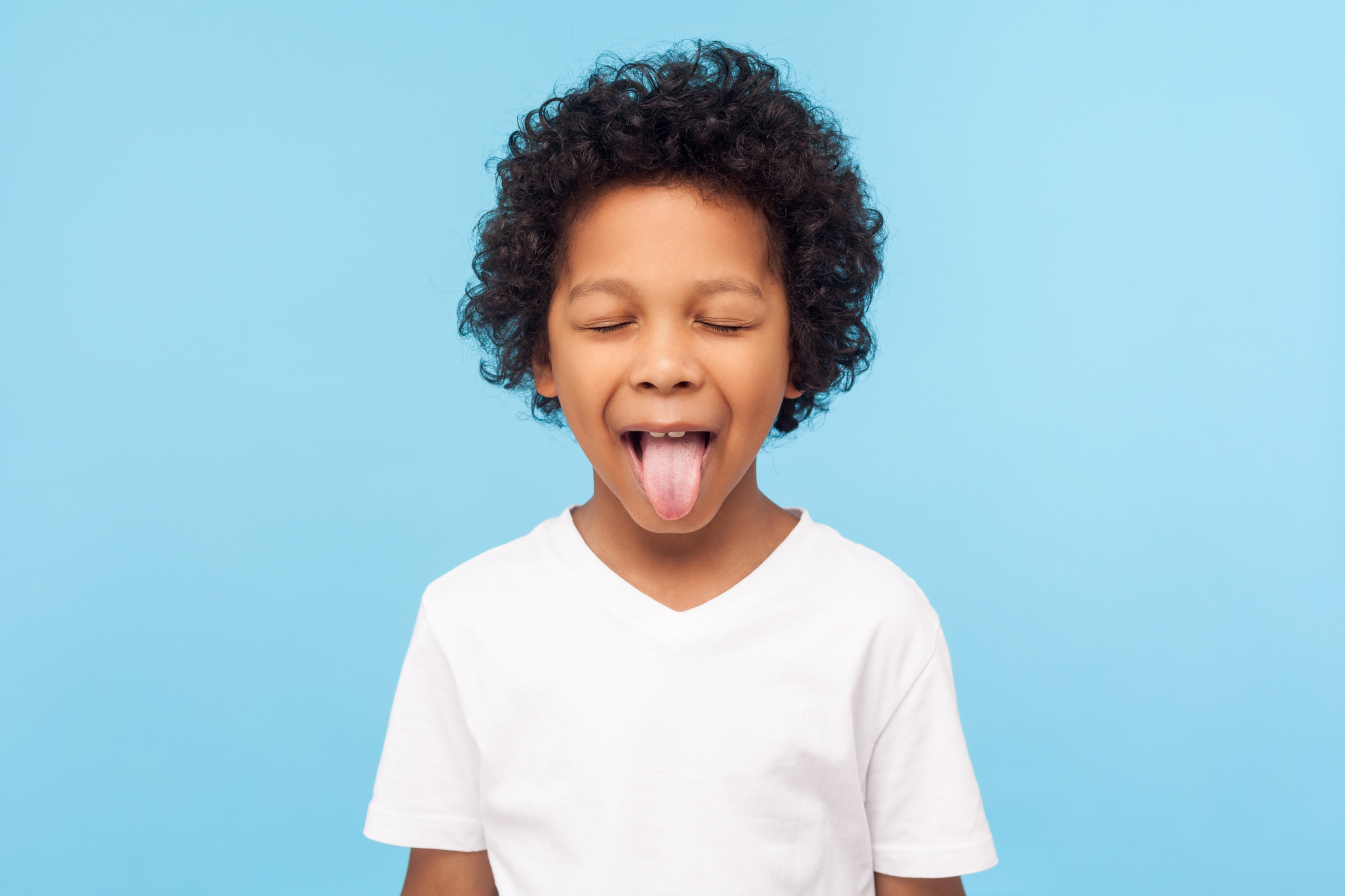 Portrait of funny naughty little boy with curly hair in T-shirt sticking out tongue and keeping eyes closed, disobedient child